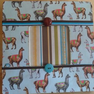 May include: A light blue picture frame with a striped paper background and a pattern of llamas. The frame has brown and blue ribbon accents and two buttons.