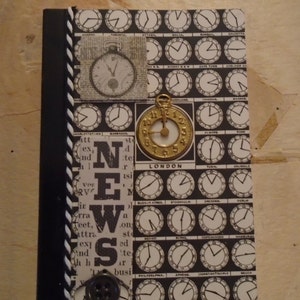 May include: A black and white notebook with a clock face design. The notebook has a black spine with a white and black braided cord. The cover has a small clock face with the word "LONDON" printed below it. The notebook also has a small clock face with a newspaper clipping behind it. The notebook has the word "NEWS" printed in large letters on the front cover.