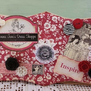 May include: A red and white floral patterned wooden sign with a white lace trim. The sign has a scalloped edge and features a white and pink label that says "Emma Jane's Dress Shoppe". The sign also has a white label that says "Inspire".