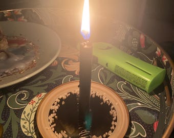 Könnte beinhalten: Eine schwarze Kerze brennt auf einer kleinen, runden, goldgeränderten Schale, die mit weißen Kristallen gefüllt ist. Die Kerze ist angezündet und die Flamme erzeugt ein warmes Leuchten.