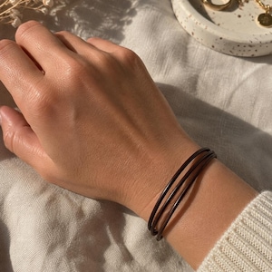 May include: A dark brown triple-strand leather bracelet worn on the wrist. The bracelet consists of three thin, smooth, rounded leather cords that wrap around the wrist, creating a layered appearance with a simple and clean design aesthetic.