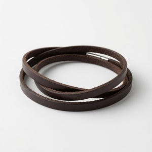 Könnte beinhalten: Ein dunkelbraunes Leder-Wickelarmband mit einem glatten, flachen Riemendesign, das sich mehrfach um das Handgelenk legt. Das Armband wird mit einem kleinen, zylindrischen, silberfarbenen Metallverschluss gesichert, der die beiden Enden verbindet.
