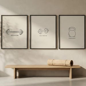 May include: Three framed prints with black line drawings of gym equipment: a barbell, a dumbbell, and a kettlebell. The prints are labelled "Discipline", "Daily Practice", and "Consistency". A rolled-up yoga mat rests on a wooden bench.