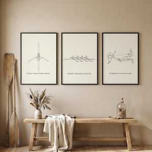May include: Three framed rowing-themed art prints with black frames. The prints feature line drawings of rowing equipment and rowers, with motivational text: "Row Your Own Race," "Every Stroke Counts," and "Strength in Rhythm." A wooden bench and oars are in the foreground.