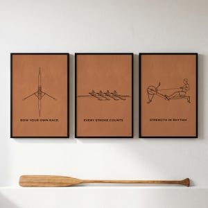 May include: Three framed art prints with brown backgrounds and black line drawings of rowing equipment and rowers. The prints have the phrases "ROW YOUR OWN RACE", "EVERY STROKE COUNTS", and "STRENGTH IN RHYTHM". A wooden oar rests below.