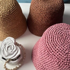 Pode incluir: Três chapéus de balde em croché nas cores castanho claro, castanho médio e rosa seco, cada um com um padrão de pontos circulares densos. Ao lado dos chapéus, encontra-se um recipiente decorativo branco em forma de coração com uma rosa esculpida e pérolas.