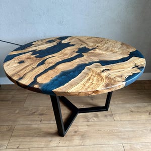 Könnte beinhalten: Runder Couchtisch aus Holz mit einzigartigem Design. Die Tischplatte hat ein natürliches Holzmaserungsmuster mit blauen Harzakzenten. Der Tisch hat ein schwarzes Metallgestell. Der Tisch hat einen Durchmesser von ca. 76 cm.