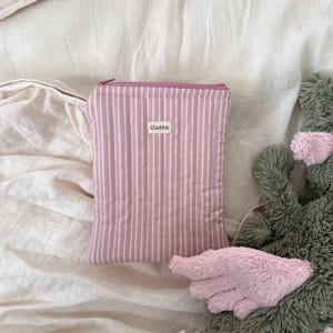 Op de afbeelding: Een rechthoekig tasje met rits voorzien van een verticaal gestreept patroon in mauve en crèmetinten. Een klein wit rechthoekig stoffen label met de zwarte tekst Catta is in het midden aan de voorzijde bevestigd. Het tasje heeft een bijpassende rits.