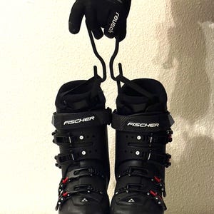 Puede incluir: Par de botas de esquí Fischer negras con detalles rojos en las hebillas y suelas. Las botas están sujetas por un guante negro. El logotipo de Fischer es visible en la parte delantera de cada bota.
