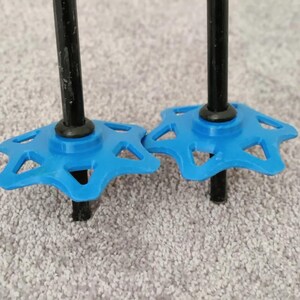 Op de afbeelding: Close-up van twee zwarte skistokken met helderblauwe stervormige manden. De manden zijn ontworpen met uitgesneden vormen. De stokken staan tegen een neutrale, getextureerde achtergrond.