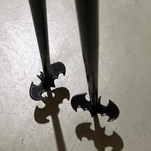 Puede incluir: Dos objetos decorativos negros con forma de murciélago y largos tallos cilíndricos. Las alas del murciélago están extendidas y los objetos proyectan sombras con forma de murciélago. Accesorios decorativos.