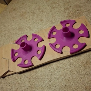 Op de afbeelding: Twee magenta cirkelvormige objecten met een centrale cilinder en meerdere cirkelvormige uitsparingen, rustend op een stuk onafgewerkt hout. De objecten lijken onderdelen te zijn van een groter item, mogelijk voor een knutsel- of doe-het-zelf project.