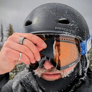 Op de afbeelding: Een zwarte skihelm met het woord "PRO-TEC" aan de zijkant. De persoon draagt een oranje getinte skibril met het merk "NFX" zichtbaar. De bril is bedekt met sneeuw. De persoon draagt twee ringen.