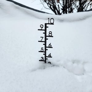 May include: A black metal snow gauge with numbers from 3 to 10, measuring the depth of snow. The gauge is placed in a snow-covered area, with the numbers clearly visible against the white background. The gauge is made of black metal.