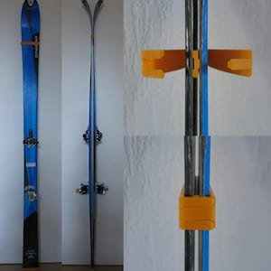 May include: A pair of blue and black skis with orange ski holders mounted on a white wall. The skis have black bindings and the brand name "Dynastar" is visible. The ski holders are made of orange plastic.