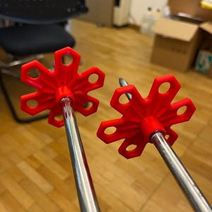 Puede incluir: Dos bastones de esquí con rosetas rojas brillantes en forma de estrella. Las rosetas tienen seis agujeros hexagonales. Los bastones son plateados y parecen ser de metal. El fondo es un suelo de madera marrón claro.