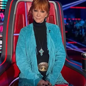 May include: A woman in a turquoise fringed jacket, black turtleneck, and a cross pendant necklace. She is seated in a red chair with a black and red design. A gold belt buckle is visible. The background is blurred.