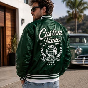 May include: A dark green satin varsity jacket featuring white striped ribbed trim on the collar, cuffs, and hem. The back displays white embroidered text reading Custom Name, a globe graphic, the words PRINT DESIGN, EST. 2024, and five stars.