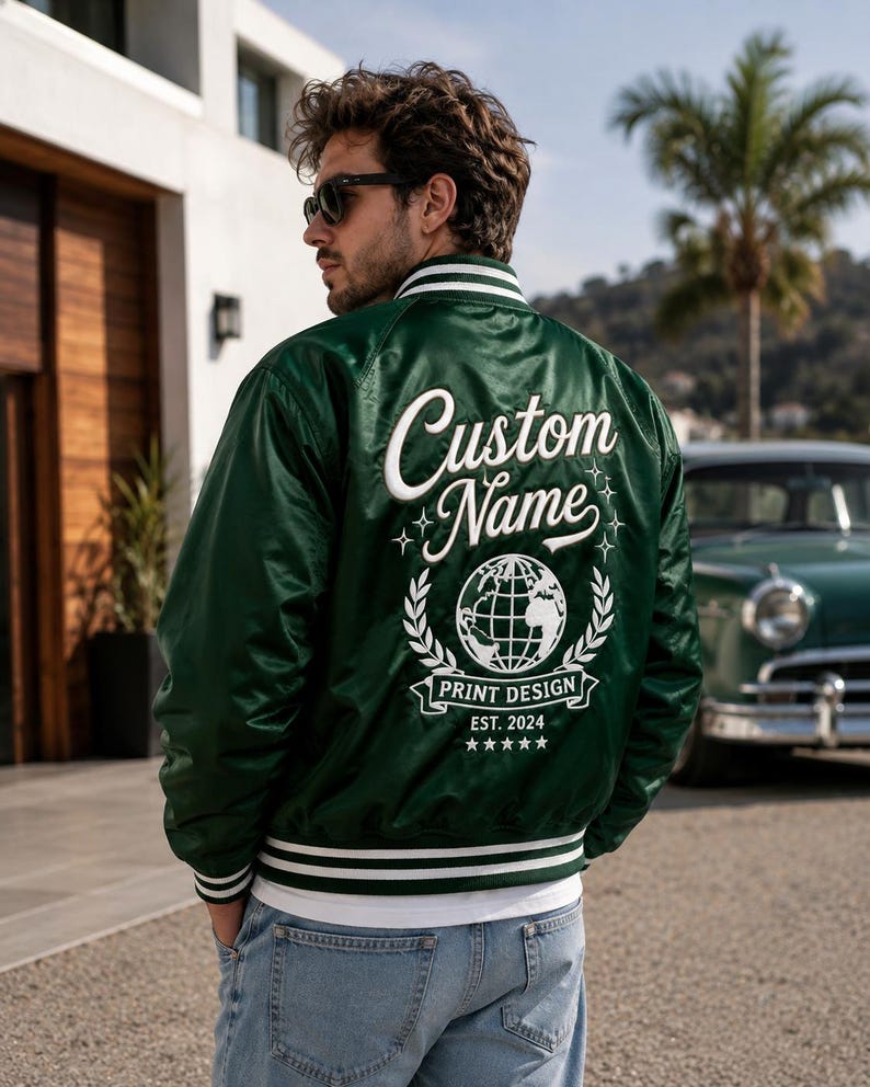 May include: A dark green satin varsity jacket featuring white striped ribbed trim on the collar, cuffs, and hem. The back displays white embroidered text reading Custom Name, a globe graphic, the words PRINT DESIGN, EST. 2024, and five stars.