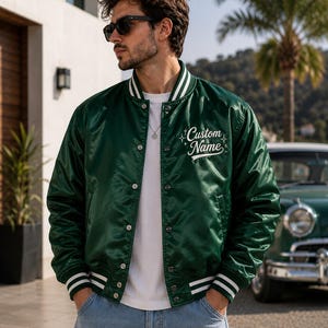 May include: A dark green satin varsity jacket featuring white striped ribbed trim on the collar, cuffs, and hem. The jacket has a snap-button front closure and the text Custom Name embroidered in white script on the left chest with star accents.