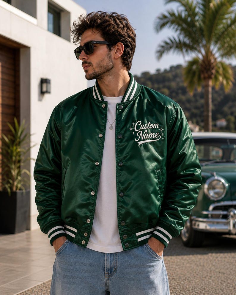 May include: A dark green satin varsity jacket featuring white striped ribbed trim on the collar, cuffs, and hem. The jacket has a snap-button front closure and the text Custom Name embroidered in white script on the left chest with star accents.