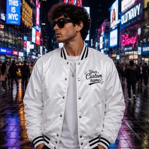 May include: A white satin varsity jacket featuring black and white striped ribbed trim on the collar and cuffs, a snap-button front closure, and the black embroidered text Your Custom name with small star accents on the left chest area.
