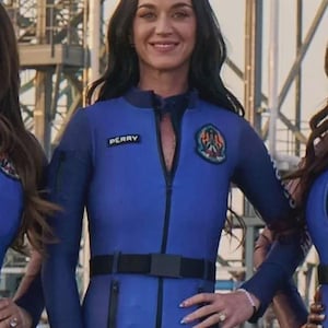 Puede incluir: Una persona con un mono azul con cinturón negro y el nombre "PERRY" en el pecho. El mono tiene un parche en el hombro derecho y una cremallera en la parte delantera. La persona sonríe y tiene el pelo oscuro.