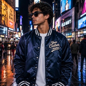May include: A navy blue satin varsity jacket with white striped ribbed trim on the collar and cuffs. The jacket features a row of silver snap buttons down the front and the embroidered text Your Custom name in white script on the left chest.