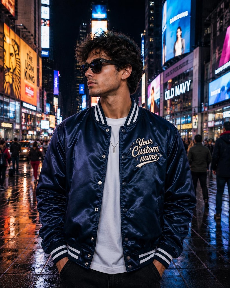 May include: A navy blue satin varsity jacket with white striped ribbed trim on the collar and cuffs. The jacket features a row of silver snap buttons down the front and the embroidered text Your Custom name in white script on the left chest.
