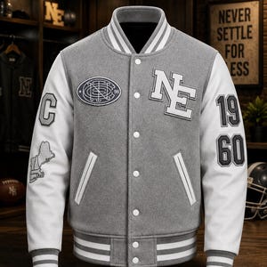 May include: A grey and white varsity jacket featuring a snap-button front, striped ribbed collar and cuffs, and various patches. The chest displays an oval emblem and the letters NE, while the sleeves feature the number 1960 and additional graphics.