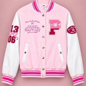 Könnte beinhalten: Eine hellrosa Collegejacke mit weißen Ärmeln, einem großen P-Aufnäher, goldenen Druckknöpfen und gestreiften Rippbündchen. Auf der Vorderseite befindet sich der Schriftzug This is Habit of Pink, während die Ärmel die Zahlen 13 und 06 zeigen.