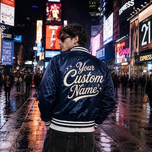 May include: A navy blue satin bomber jacket featuring white striped ribbed trim on the collar, cuffs, and hem. The back of the jacket displays the embroidered text Your Custom Name in a cursive font, accented with small star symbols in white thread.