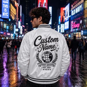 May include: A white satin varsity jacket featuring black striped ribbed trim on the collar and cuffs. The back displays black embroidered text reading Custom Name, a globe graphic, the words Print Design, Est. 2024, and five small stars.