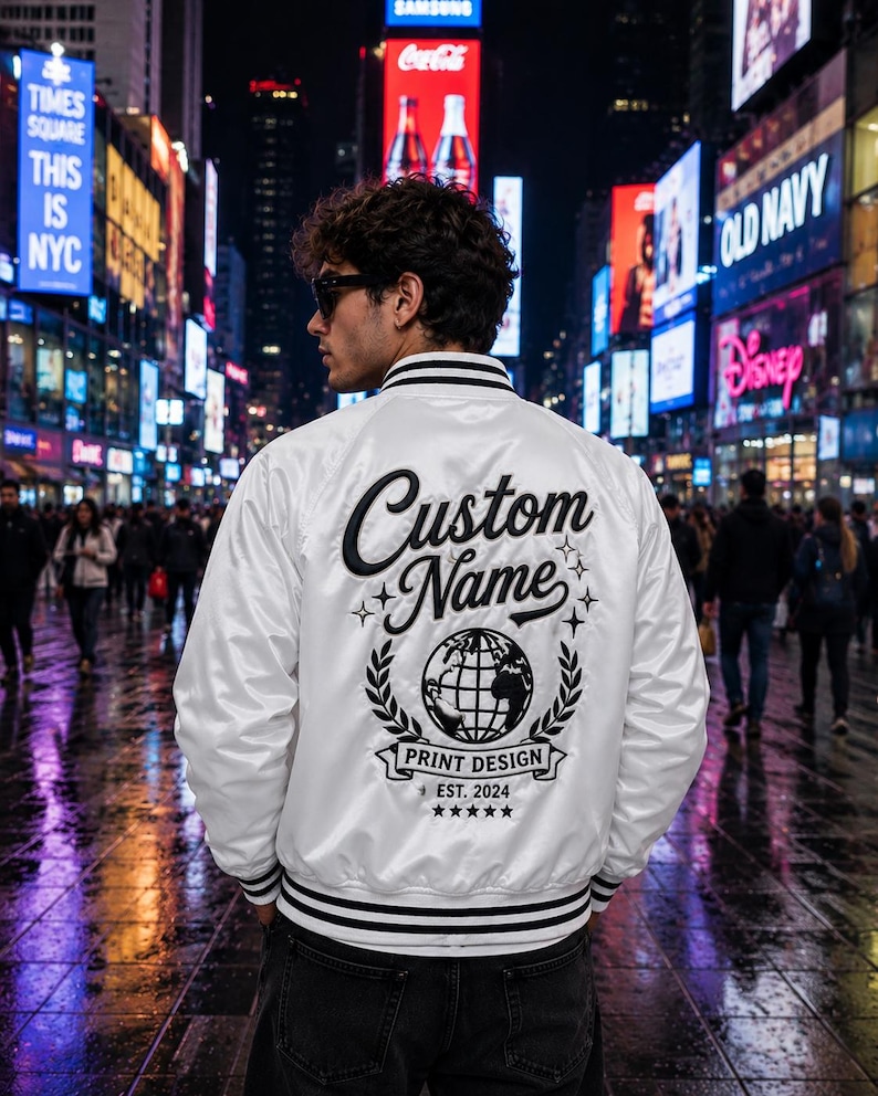 May include: A white satin varsity jacket featuring black striped ribbed trim on the collar and cuffs. The back displays black embroidered text reading Custom Name, a globe graphic, the words Print Design, Est. 2024, and five small stars.