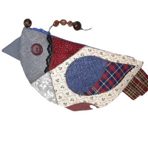May include: A whimsical bird-shaped pouch, crafted from various fabric patches in shades of gray, red, blue, and cream. The pouch features a button closure and a beaded accent. The design incorporates floral and plaid patterns.
