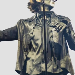 Puede incluir: Un artista con una chaqueta de cuero negra, camisa y guantes, con una máscara y pintura facial. La chaqueta está abotonada y la camisa tiene una corbata larga. El artista sostiene un micrófono.