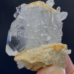 Puede incluir: Un grupo de cristales de cuarzo transparente con múltiples puntas y facetas. El cuarzo está montado sobre una matriz beige con toques de naranja y marrón. El fondo es azul oscuro.