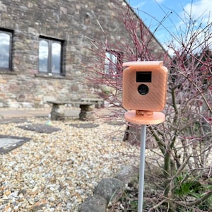 May include: A small, peach-colored security camera mounted on a gray pole. The camera is in a garden setting with a stone wall and a small tree in the background.
