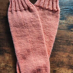 May include: A pair of coral-colored knitted socks. The socks are made of a soft, textured yarn and feature a ribbed cuff. The socks are laying on a wooden surface. The socks appear to be handmade.