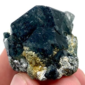 以下が含まれることがあります： 暗い青緑色の鉱物標本で、鋭く角張った結晶構造をしています。鉱物は、黄色と白のヒントがある、より明るい色の岩のマトリックス上にあります。 全体的な外観は粗く自然で、石の生の美しさを表現しています。