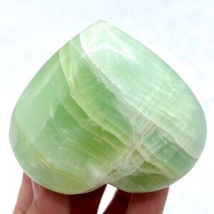 May include: A heart-shaped, polished green calcite stone. The stone has a smooth surface with visible layers and variations in colour, ranging from light to medium green. The heart is a solid, opaque object.