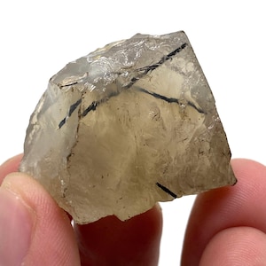 以下が含まれることがあります： 黒トルマリンのインクルージョンが入った、生の半透明な水晶。 水晶は薄茶色で、白い背景に対して2本の指で持たれています。 天然石は、粗く、質感のある表面を持っています。