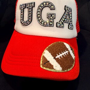 May include: A red and white trucker hat featuring the letters UGA in large, iridescent rhinestone-encrusted block letters on the front panel. A brown and white felt football applique with gold trim is stitched onto the red brim of the cap.
