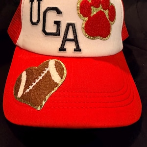 May include: A red and white trucker hat featuring the letters UGA in black, a red paw print patch with a gold glitter border, and a brown football-shaped patch with white laces on the brim, all displayed on a solid red background.