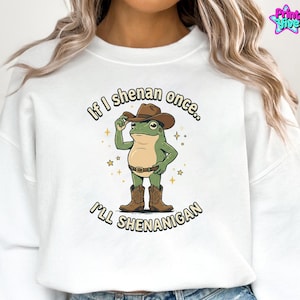 May include: White sweatshirt with a cartoon frog wearing a cowboy hat, boots, and belt. The frog is holding up the hat with one hand. Text on the sweatshirt reads "If I shenan once... I'll shenanigan."