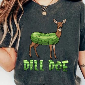 Puede incluir: Camiseta gris oscuro con un ciervo de dibujos animados con cuerpo de pepinillo. El texto verde "DILL DOE" está impreso debajo. La camiseta es de estilo casual.