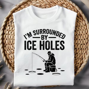 Peut inclure: T-shirt blanc avec le texte "I'M SURROUNDED BY ICE HOLES" en noir. Une silhouette noire d'une personne pêchant sur glace est en dessous du texte. Le t-shirt est présenté sur un panier en osier.