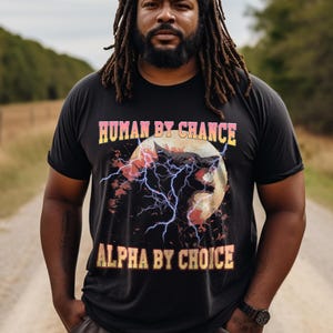 May include: Black t-shirt with the text "HUMAN BY CHANCE" and "ALPHA BY CHOICE" in yellow and pink lettering. The shirt features a wolf graphic with lightning bolts and a full moon design.