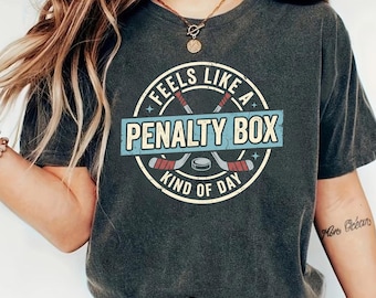May include: Dark gray t-shirt with a distressed graphic. The graphic reads "Feels Like A Penalty Box Kind of Day" in a circular design with hockey sticks and a puck. The shirt is a casual, unisex style.