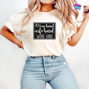 May include: Cream-colored t-shirt with the text "Mom hard wife hard WORK HARD" in a black rectangle. The shirt is paired with blue jeans. The image is well-lit, showcasing the casual style of the top.